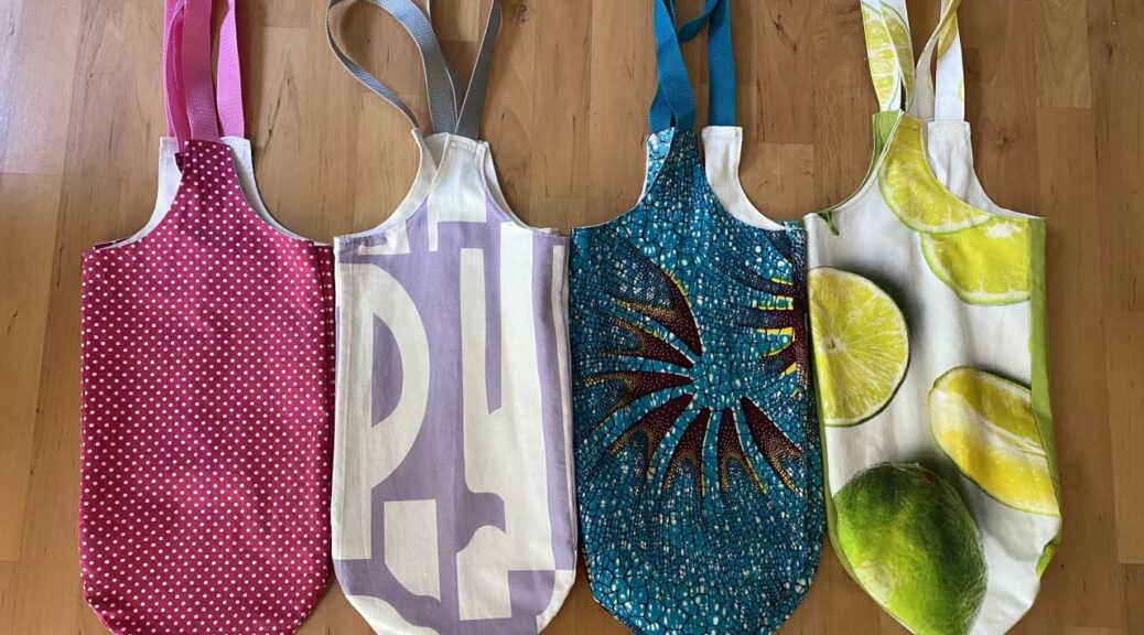 Auf einem Laminatboden liegen nebeneinander vier längs-gefaltete Taschen in folgenden Farben: rosa mit kleinen weißen Punkten und pinke Tragegurte, lila mit großem weißem Muster und graue Gurte, blau-gelb-lila gesprenkelt mit Muster und blauen Gurten, große halbierte und ganze Zitronen auf weißem Grund und grünem Stoff, Gurte aus gleichem Stoff.