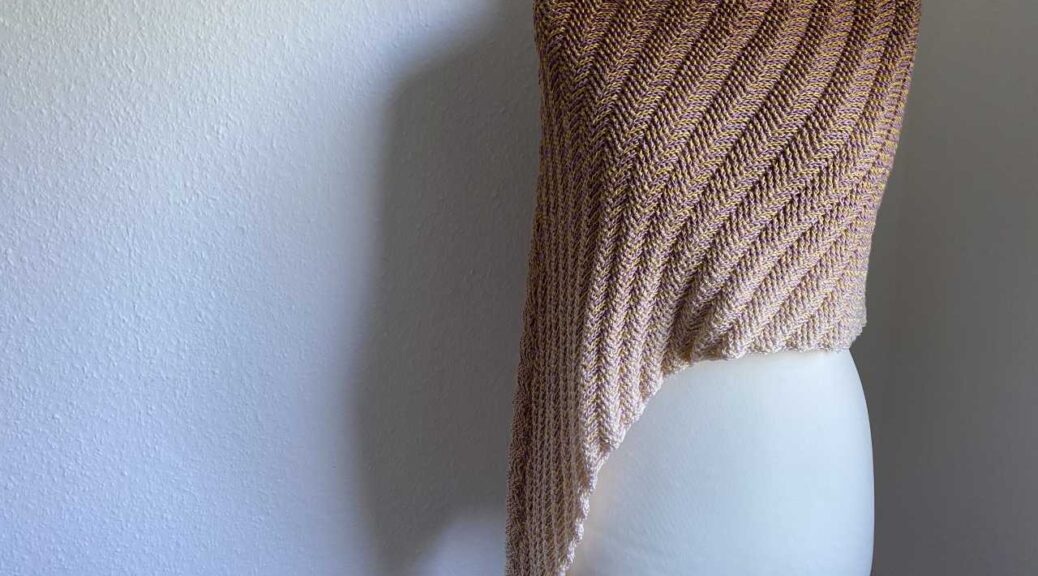 Eine Schneider Puppe steht etwas seitlich vor einer weißen Wand, die etwas im Schatten liegt. Ein großer Überwurf liegt über der Puppe, der Überwurf ist seitlich länger. Der Farbverlauf verläuft vom Hals bis über die Hüfte von dunkel bis sehr hell. Das Foto ist im Querformat aufgenommen.