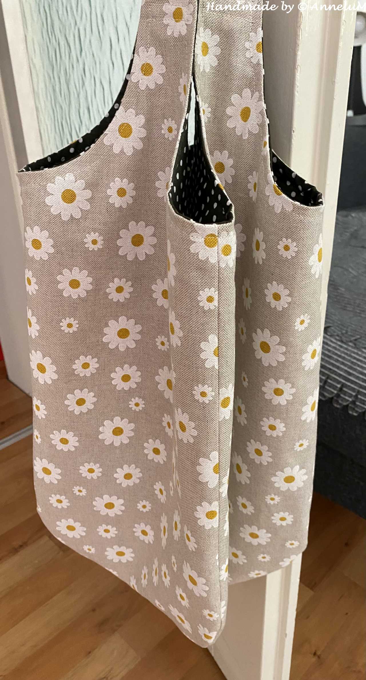 Das Bild zeigt eine handgefertigte Einkaufstasche, die an einer Tür hängt. Die Tasche ist aus einem hellbraunen Stoff mit einem Muster aus weißen Gänseblumen. Die Tasche hat zwei breite Träger. Der Innenteil der Tasche ist dunkelbraun mit weißen Punkten, was einen schönen Kontrast zum äußeren Design bildet. Die Tasche hat eine Öffnung, die leicht geöffnet ist, um den Inhalt zu zeigen. Der Boden des Raums besteht aus einem hellbraunem Laminatboden, Die Tasche wirkt robust und praktisch, ideal für Einkäufe oder den täglichen Gebrauch.