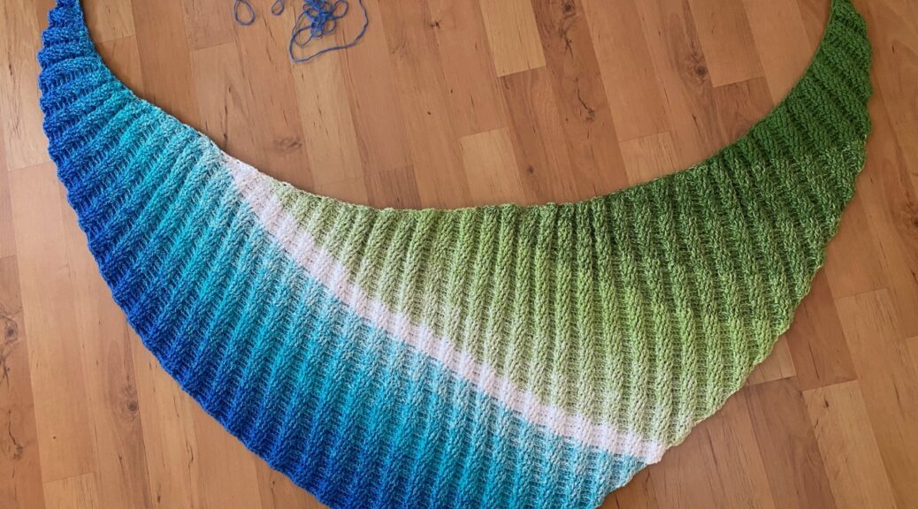 Das Bild zeigt ein gehäkeltes Dreiecktuch, das auf einem Laminatboden ausgebreitet liegt. Das Stück hat eine elegante, halbkreisförmige Form und zeigt einen sanften Farbverlauf von Blau über Türkis und Weiß bis hin zu Grün. Die Textur der Wolle weist ein rippiges Muster auf. Einige Fäden des restlichen Garns liegt unordentlich auf dem Boden. Im oberen rechten Bereich des Bildes ist der Text "Handmade by © AnneluM" zu sehen, was darauf hinweist, dass es sich um ein handgefertigtes Stück handelt.