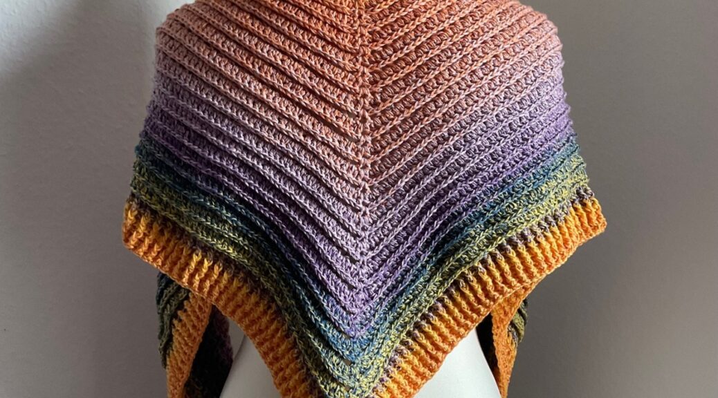 Das Bild zeigt ein buntes Häkeltuch mit Rippen, das über den Rücken einer Schneiderpuppe hängt. Diese steht vor einer weißen Wand. Das Tuch hat einen sanften Farbverlauf von innen dunkelorange in einen lila-Verlauf, dann zu grün, hellgrün und gelb. Im oberen rechten Bereich des Bildes ist der Text "Handmade by © AnneluM" zu sehen.